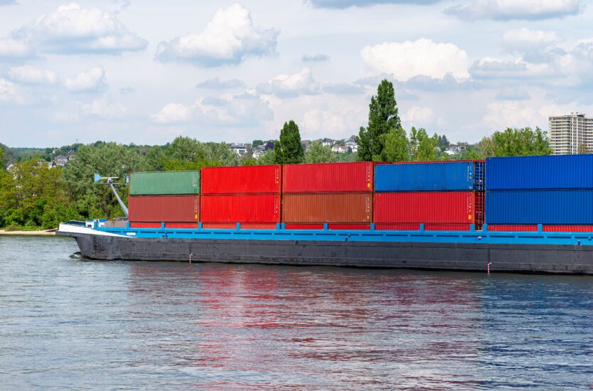 A barge carrying many containers on the Rhine in western Germany, trees and buildings in the background.